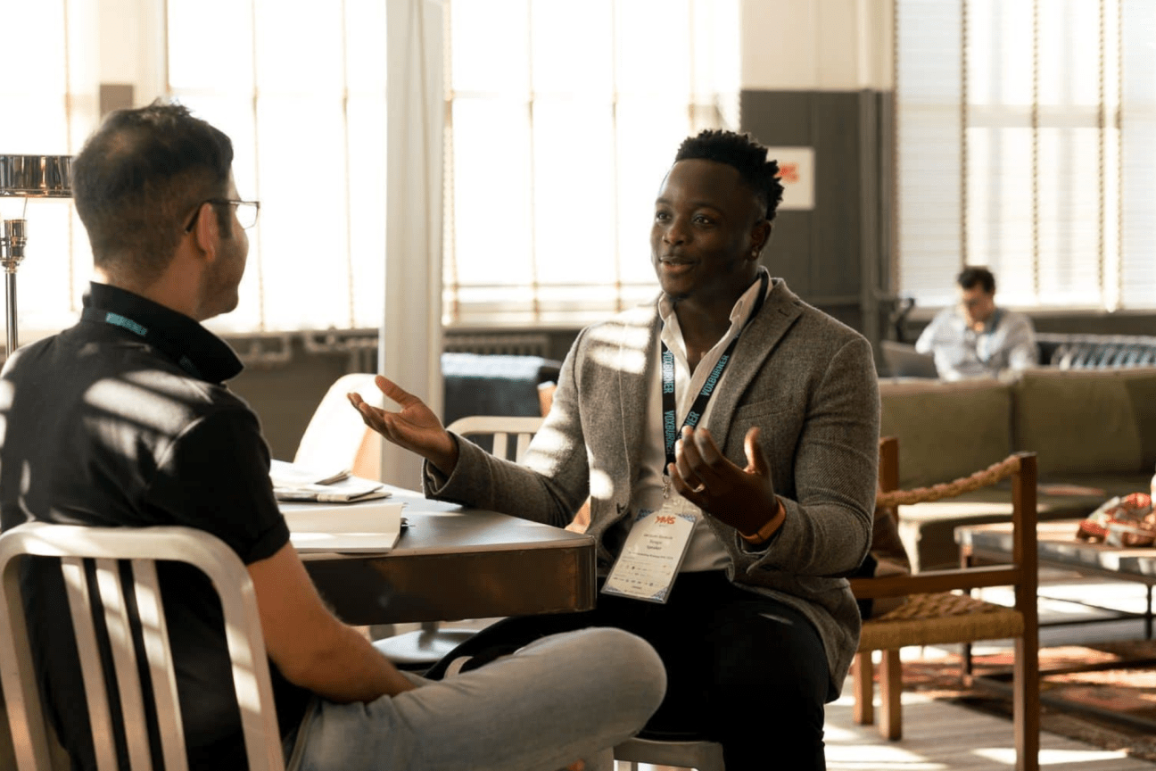 Two men sitting talking at a table. One has his back to us, the other is gesturing with his arms.