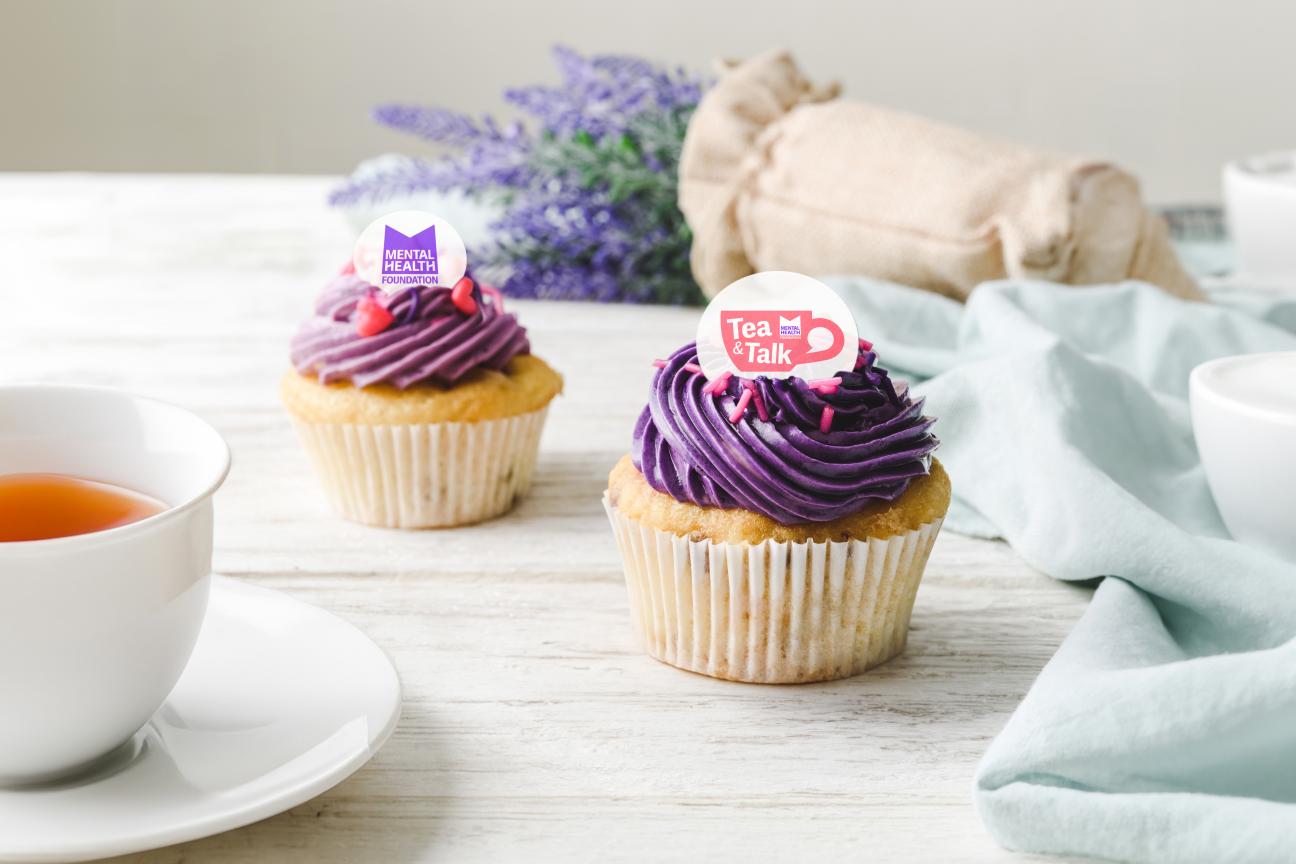 A pair of cupcakes with purple icing and circular Tea & Talk cake topper on top