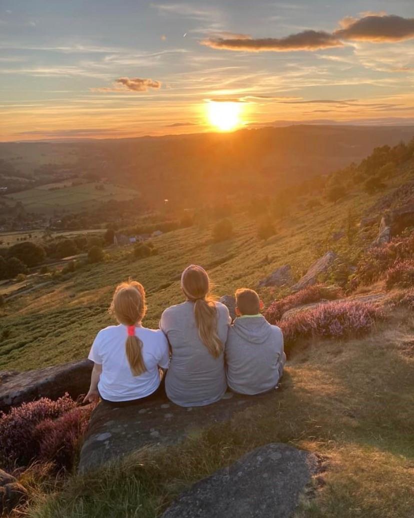 A woman is sat between two children with their backs to the camera, looking out at a sunset over green hills.