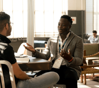 Two men sitting talking at a table. One has his back to us, the other is gesturing with his arms.