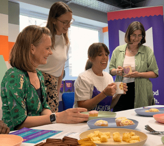 A group of smiling people sitting and standing a table with cake, biscuits and cups of tea on it