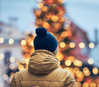 The back of a person wearing a winter hat and coat, facing towards a street with festive lights.