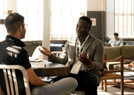 Two men sitting talking at a table. One has his back to us, the other is gesturing with his arms.