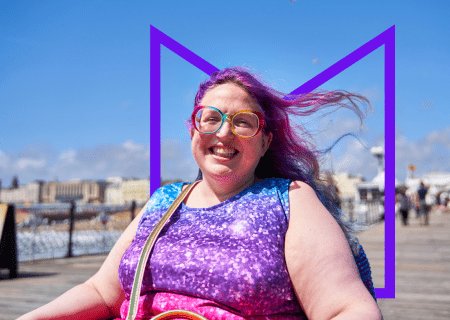 A woman in a wheelchair on a pier on a sunny day. She's smiling at the camera and her purple-dyed hair is blowing in the wind.